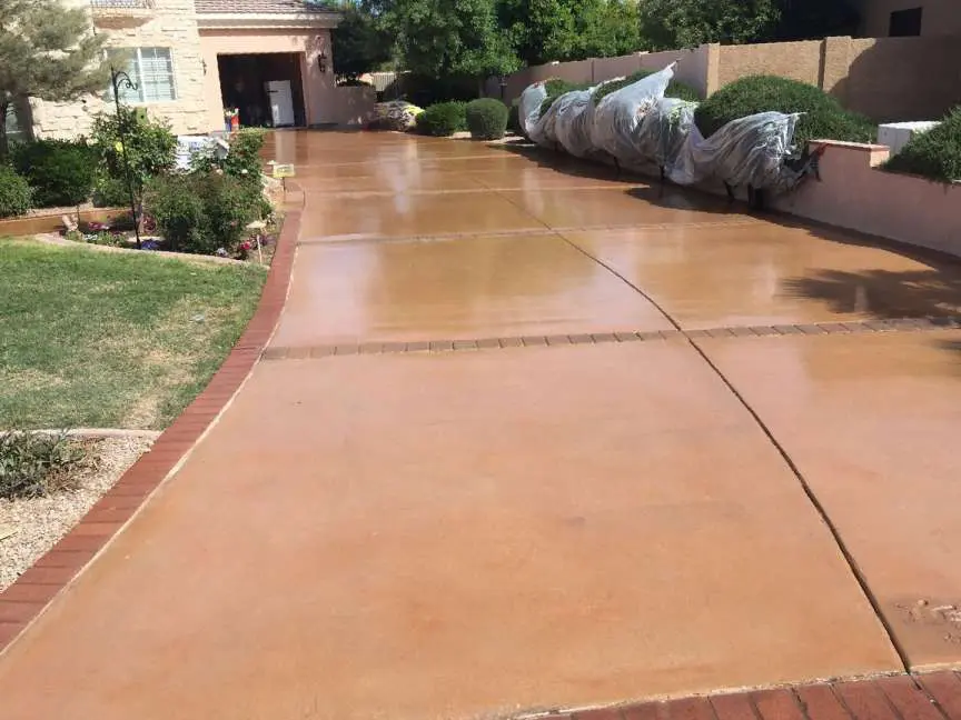 A wide, freshly cleaned driveway in front of a suburban house in Miami, FL. The driveway, expertly crafted by a decorative concrete contractor, is bordered by brick and has a well-maintained grassy area on the left. Along the right side, bushes are covered with plastic tarps.