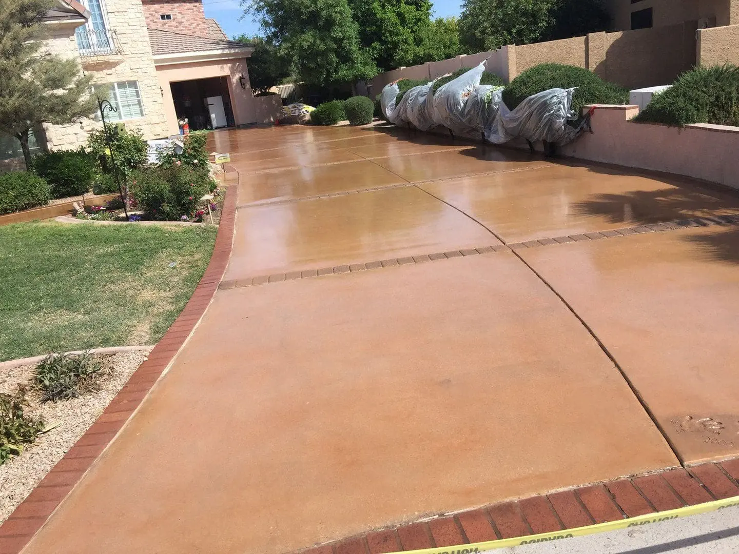 A long driveway with a freshly washed and finished concrete surface, courtesy of top-tier Concrete Contractors Miami, leads to a residential garage. The driveway is bordered by red bricks and surrounded by garden beds and neatly trimmed bushes. A garden hose lays on the left, with wrapped objects on the right side.