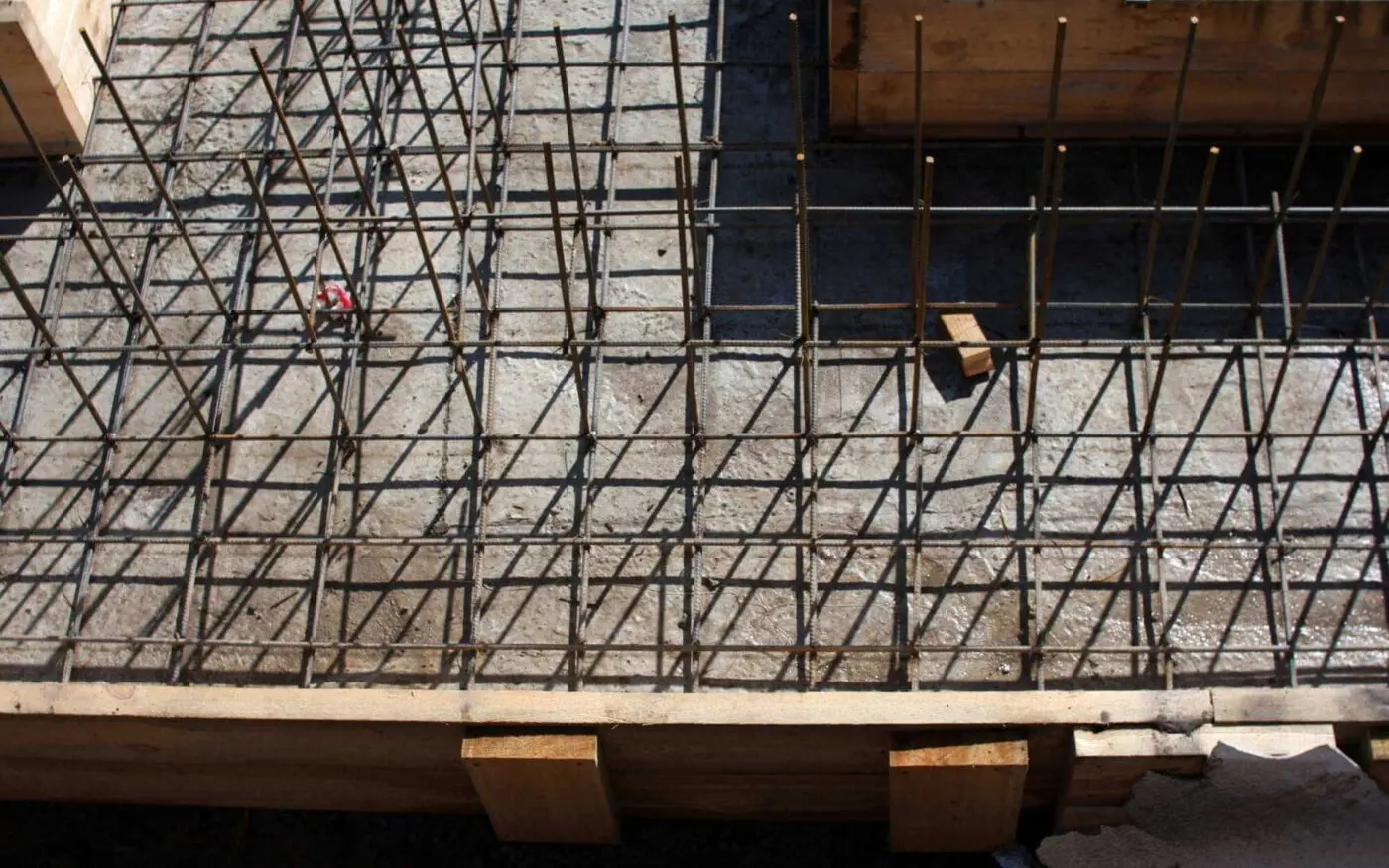 An overhead view of a Miami concrete construction site shows a concrete slab reinforced with a grid of steel rebar. Wooden planks line the edges of the slab, and a small red object sits on the left side. Shadows from the rebar cast a geometric pattern on this decorative concrete surface.