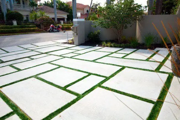 A modern driveway in Miami, FL features large, rectangular concrete slabs separated by strips of green grass. Designed by a decorative concrete contractor, the geometric layout contrasts the hard concrete with the soft grass. A residential street and houses are visible in the background.