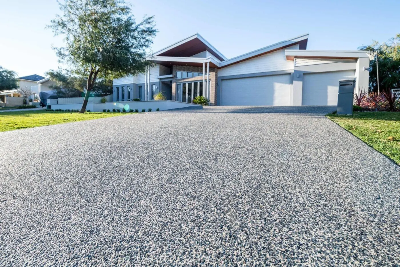 A modern, single-story house with a wide driveway, thanks to Concrete Contractors Miami, leading up to a three-car garage. The home features large windows, a sleek roof design, and minimalistic landscaping with grass and trees. The sky is clear and blue, suggesting a sunny day.