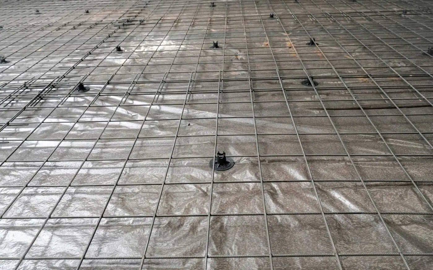 A close-up view of a construction site floor in Miami shows a grid of steel reinforcement bars placed over a plastic sheet, supported by small plastic spacers. The steel bars form a pattern of squares on the surface, an essential step before pouring concrete slabs often associated with Dade Decorative Concrete standards.