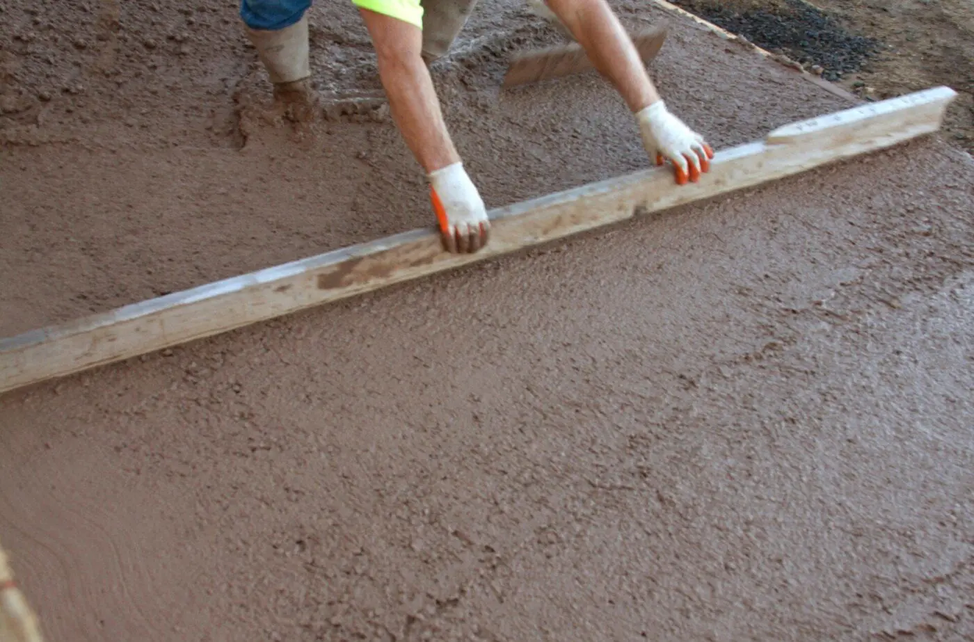 A construction worker in a high-visibility shirt and gloves uses a long wooden plank to level freshly poured decorative concrete on the ground. The worker’s legs and arms are visible, with the concrete surface appearing smooth and wet.