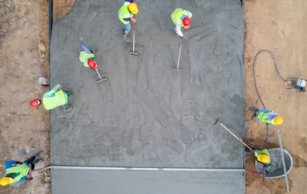 Aerial view of construction workers wearing high-visibility vests and helmets, spreading and smoothing wet decorative concrete on a large rectangular slab. They are using different tools to ensure an even surface, with equipment and materials visible on the surrounding ground.