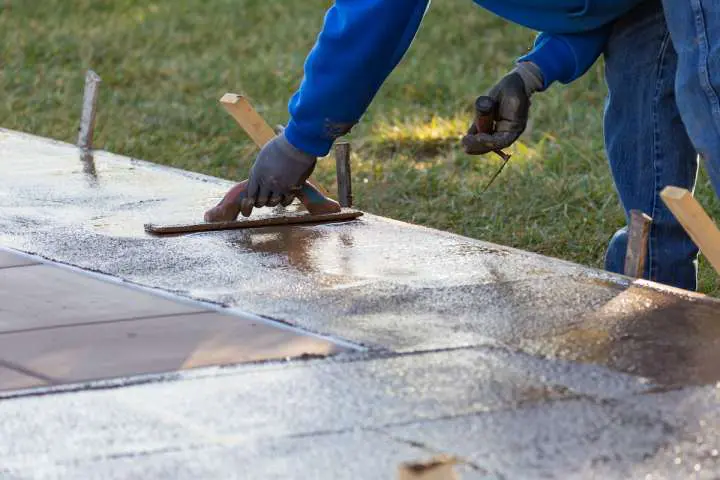 A person wearing a blue hoodie and gloves is smoothing wet decorative concrete with a trowel. Wooden stakes mark the perimeter, and nearby grass is visible. The work is being done outdoors on a sunny day in Coral Gables, FL.