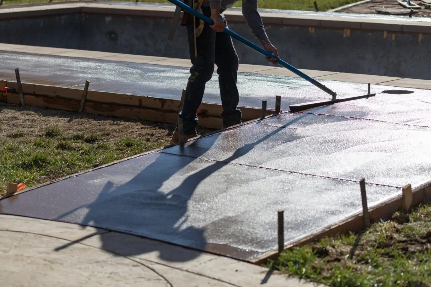 A construction worker employed by concrete contractors in Hialeah, FL, uses a long-handled tool to smooth freshly poured concrete on a walkway. The scene unfolds outdoors under bright sunlight, with shadows cast on the damp cement. Wood forms outline the area, and other construction materials are visible.