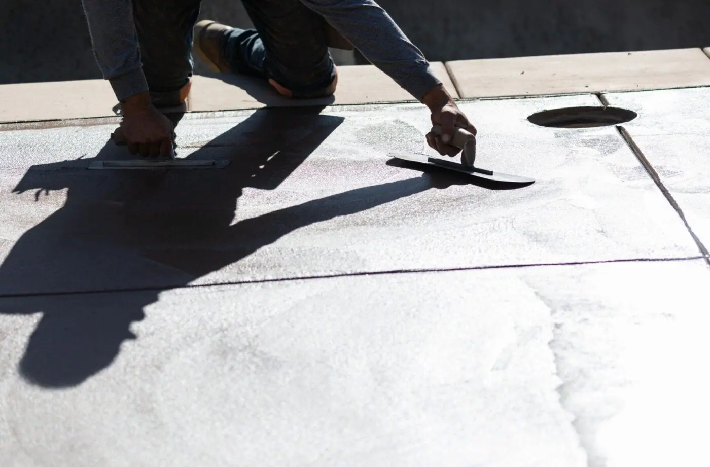 A person troweling smooth concrete with tools, casting a shadow over the slab. The individual, representing Dade Decorative Concrete, wears a long-sleeved shirt and kneels to apply even pressure, ensuring a flat and consistent surface. The scene is lit with sunlight, creating strong contrast and shadows.