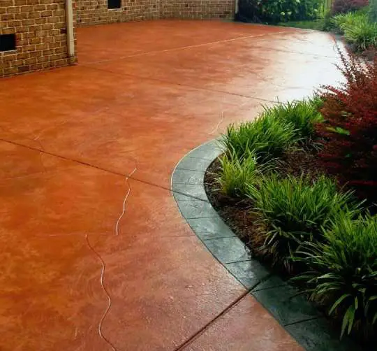 A smooth, reddish-brown concrete patio curves alongside a garden bed. The garden features green ornamental grasses and other shrubs, bordered by a stone trim. The patio, crafted by skilled concrete contractors in Miami, is next to a brick foundation wall.