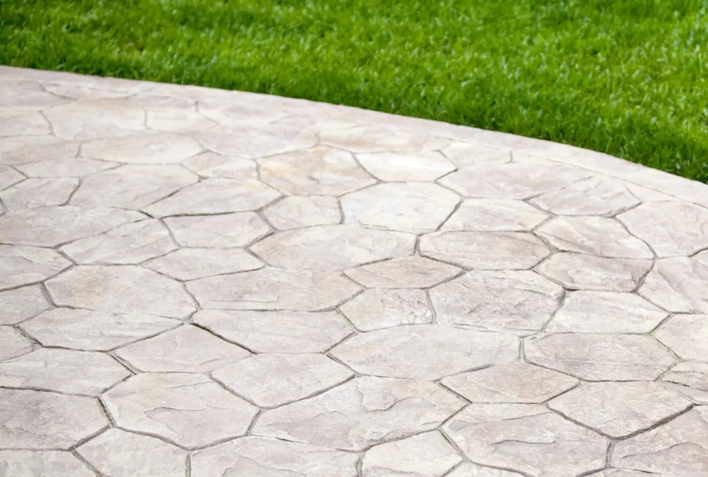 A close-up view of a stone patio with a textured, irregular-shaped pattern, typical of concrete patios in Miami FL. The patio lies adjacent to a lush, green lawn, providing a contrast in colors and textures. The scene is outdoors, suggesting a garden or backyard setting designed by a decorative concrete contractor.