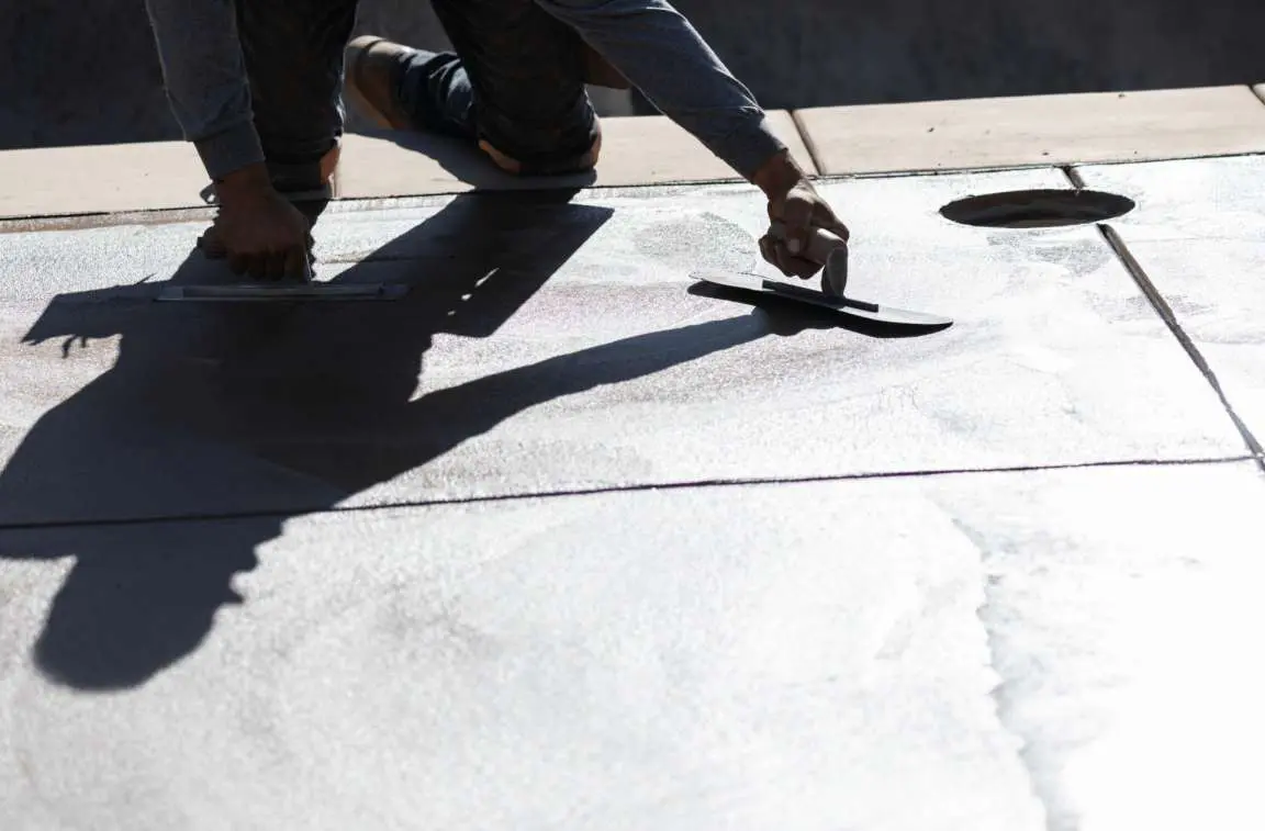 A person is kneeling on the ground, using a trowel to smooth out concrete at a busy construction site. The shadow of their outstretched arm is visible on the surface. For top-notch concrete services Miami-Dade residents trust, this scene exemplifies the expertise of Concrete Contractors Miami.