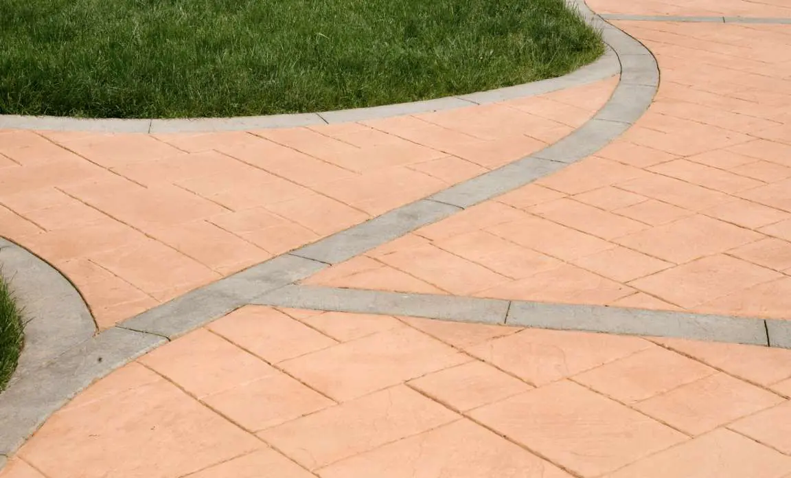 A neatly designed outdoor pathway made of reddish-orange tiles with light gray borders curves through a green grass lawn in Miami, FL. The pathway, crafted by expert concrete contractors, divides into two directions, creating a visually appealing geometric pattern. The grass is vibrant and well-maintained.