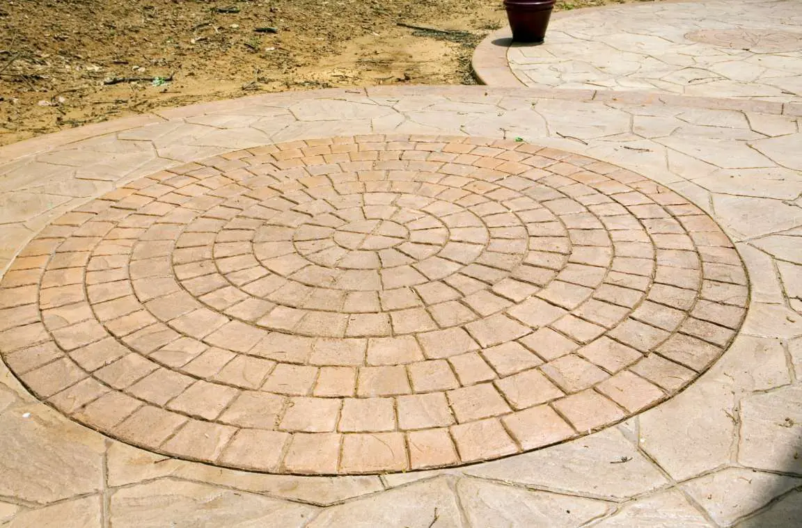 A circular brick pattern is set in the middle of a beige stone patio. The brick design radiates outward from the center, creating a geometric look. A small brown planter with a plant is positioned near the upper edge of the photo on a similar patio surface by Concrete Contractors Miami.