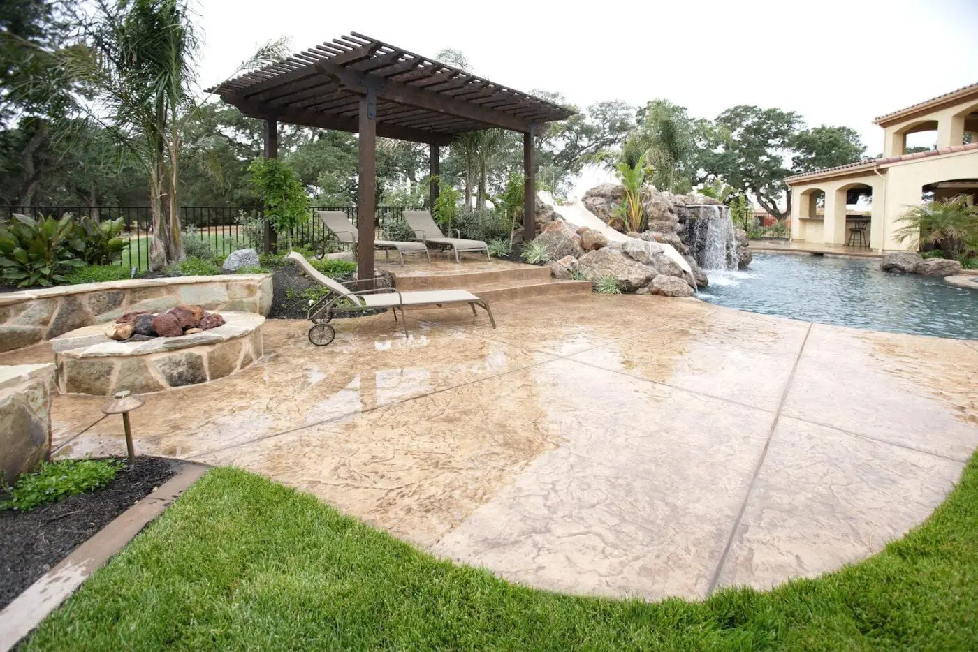 A luxurious backyard in Miami, FL features a stamped concrete patio with two reclining loungers and a shaded pergola. The pool area includes a waterfall and is surrounded by lush greenery and trees. A stone fire pit is near the pergola, while a large house stands majestically in the background, crafted by a top decorative concrete contractor.