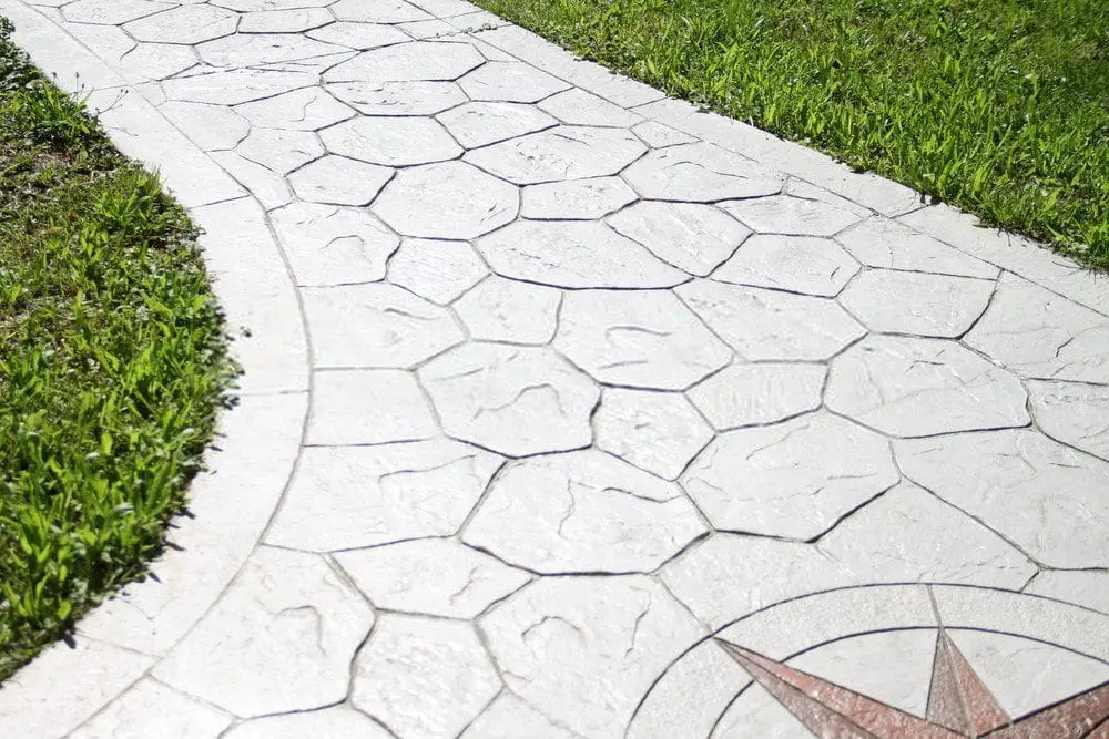 A stone pathway with hexagonal-shaped stones and fossil-like imprints of leaves and bones winds through a grassy area, offering a driveway facelift. A compass rose is embedded in the lower section of the pathway, showcasing modern concrete solutions.