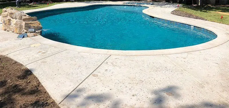 A kidney-shaped swimming pool with clear blue water is surrounded by a concrete deck, reminiscent of Miami FL's stylish outdoor spaces. Rocks adorn one end, with patches of grass and dirt around the edge. Trees and shrubs provide a serene backdrop. Contact us for Free Estimates on Concrete Pool Deck Installations.