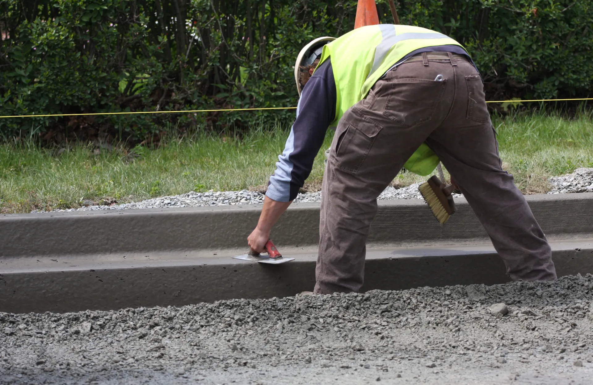 A construction worker from Dade Decorative Concrete in Coral Gables, FL, wearing a high-visibility vest and a hard hat, smooths out wet concrete on a sidewalk using a trowel. The worker is bent over, concentrating on their task. There are trees and a grassy area in the background, ideal for decorative concrete projects.