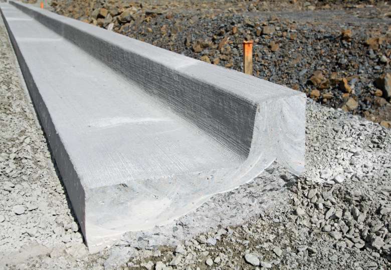 Concrete drainage channel under construction, running along a gravel path with exposed rocky earth on the side. Orange guide markers are seen in the background, indicating the alignment and depth. Concrete Contractors Miami offer expertise and precision for such projects, ensuring quality results.