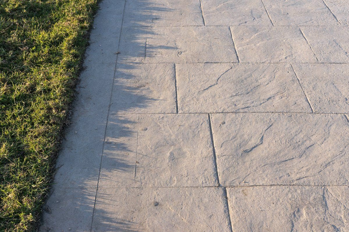 A close-up of a paved walkway with neatly cut grass on the left side in Miami, FL. The walkway, reminiscent of decorative concrete driveways, features large, textured stone slabs. Shadows of the grass blades extend across the edge of the pavement, creating a natural pattern.
