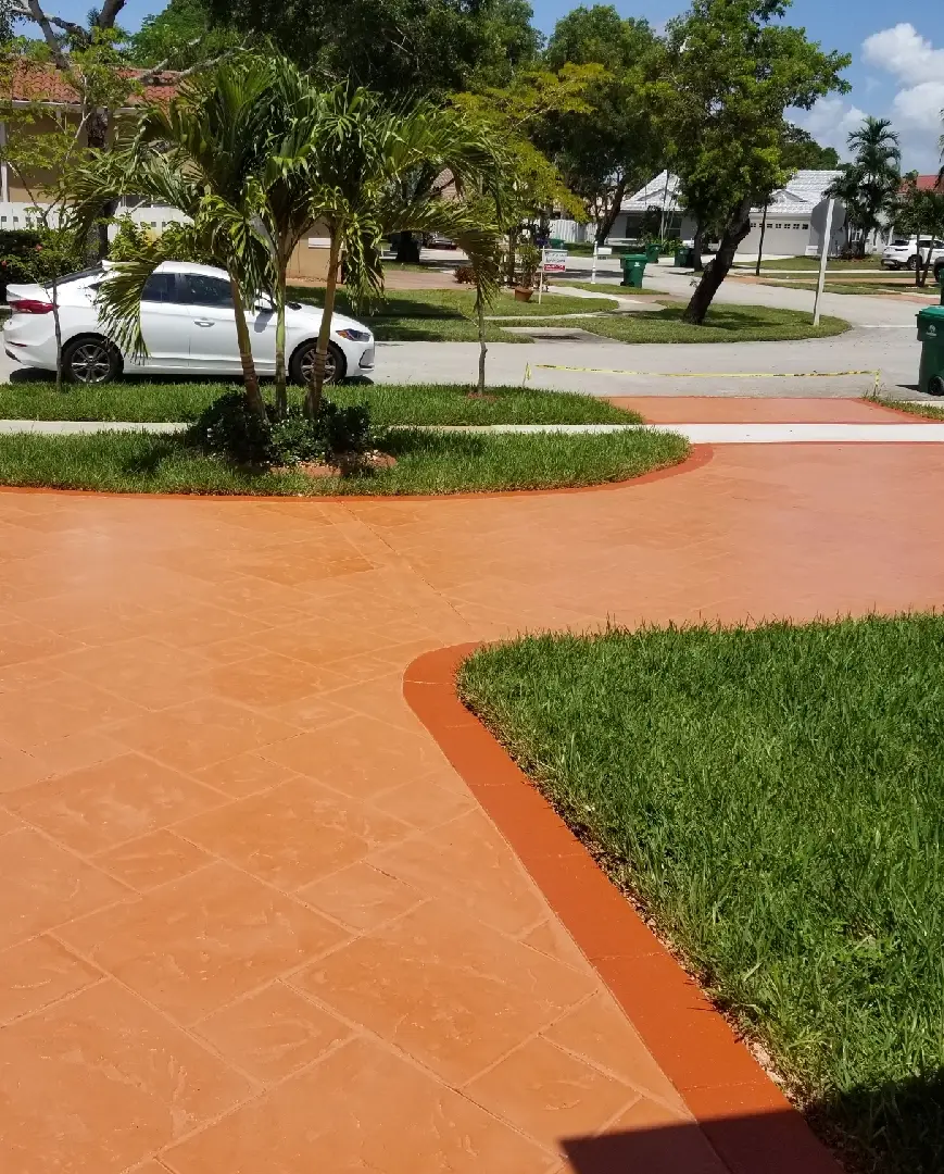 A long driveway with a freshly washed and finished concrete surface, courtesy of top-tier Concrete Contractors Miami, leads to a residential garage. The driveway is bordered by red bricks and surrounded by garden beds and neatly trimmed bushes. A garden hose lays on the left, with wrapped objects on the right side.